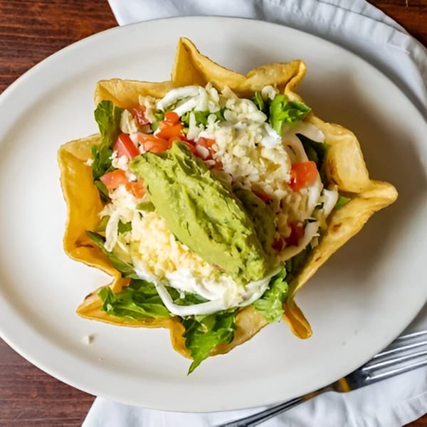 A taco salad in a crispy tortilla bowl, topped with guacamole, lettuce, tomatoes, and cheese.